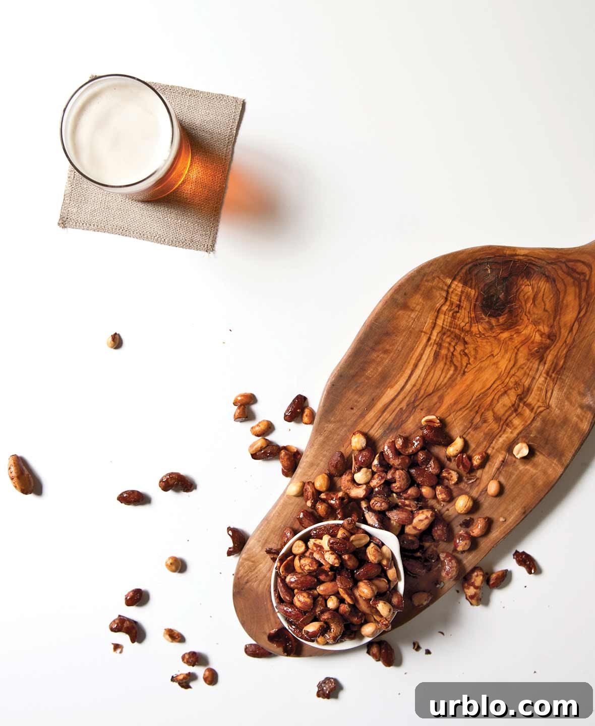 An overflowing bowl of beer nuts on a wooden board with a glass of beer on a napkin beside it.
