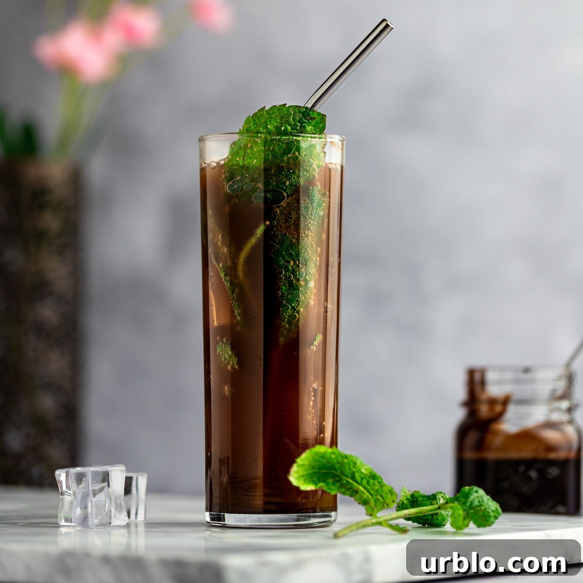 Up close photo of a chocolate mojito, with a metal straw and garnished with fresh mint leaves, on a marble board, next to mint leaves and ice cubes, with pink flowers behind.