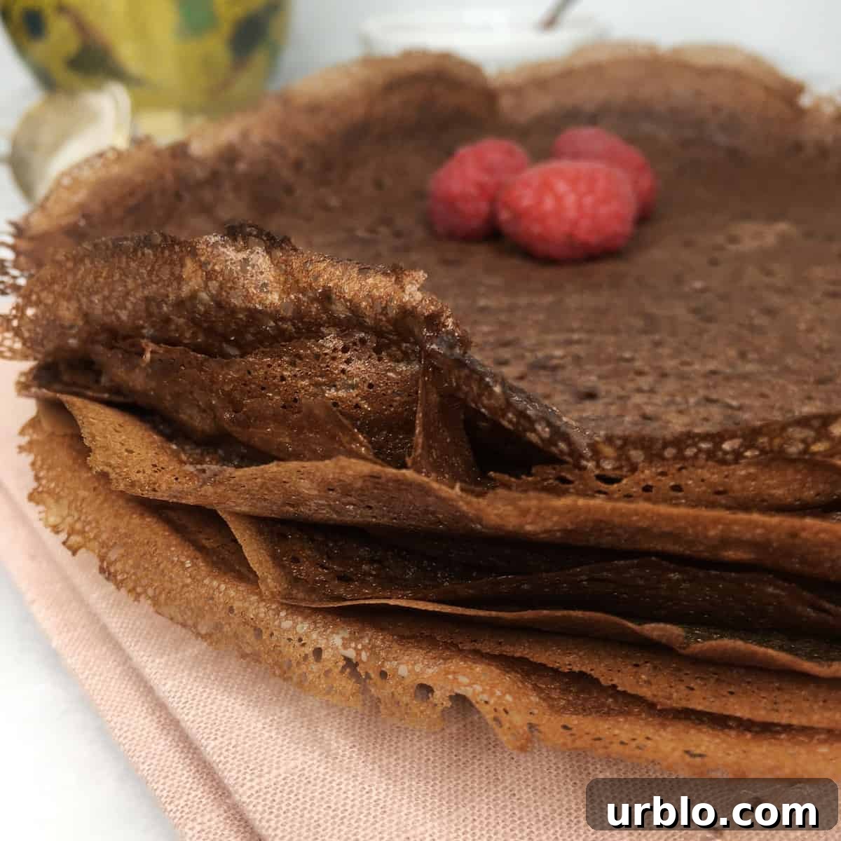 A batch of chocolate crepes with a pink tea towel and a cup in the background