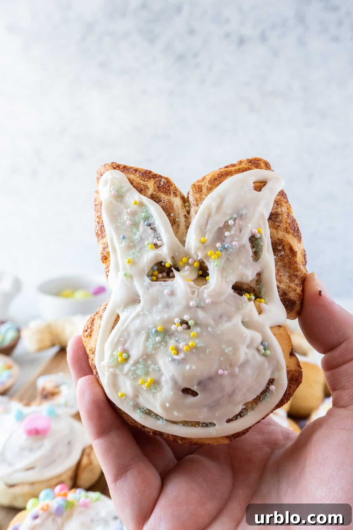 bunny cinnamon roll frosted and decorated with sprinkles being held in hand.