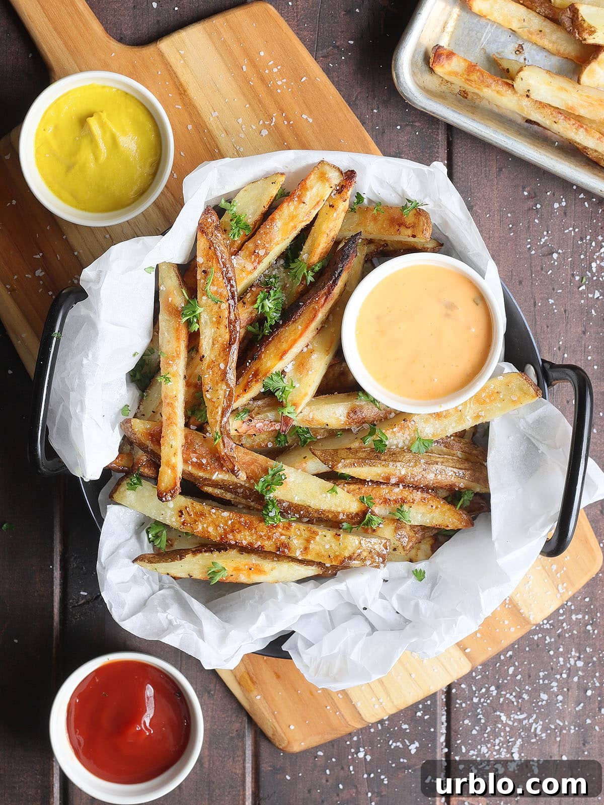 Oven baked French fries in a parchment paper lined dish garnished with salt and chopped fresh parsley.