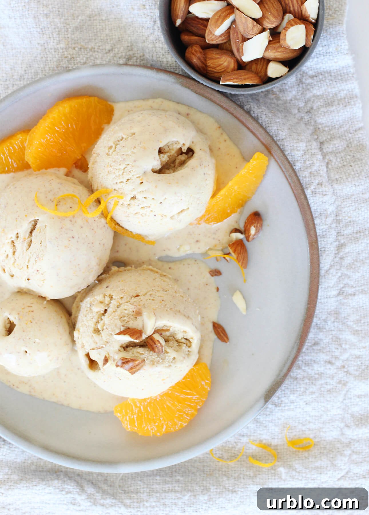 Scoops of orange almond gelato are garnished with sliced almonds and orange segments on a plate. A small bowl filled with almonds is placed beside the plate, all resting on a textured beige cloth. // FoodNouveau.com