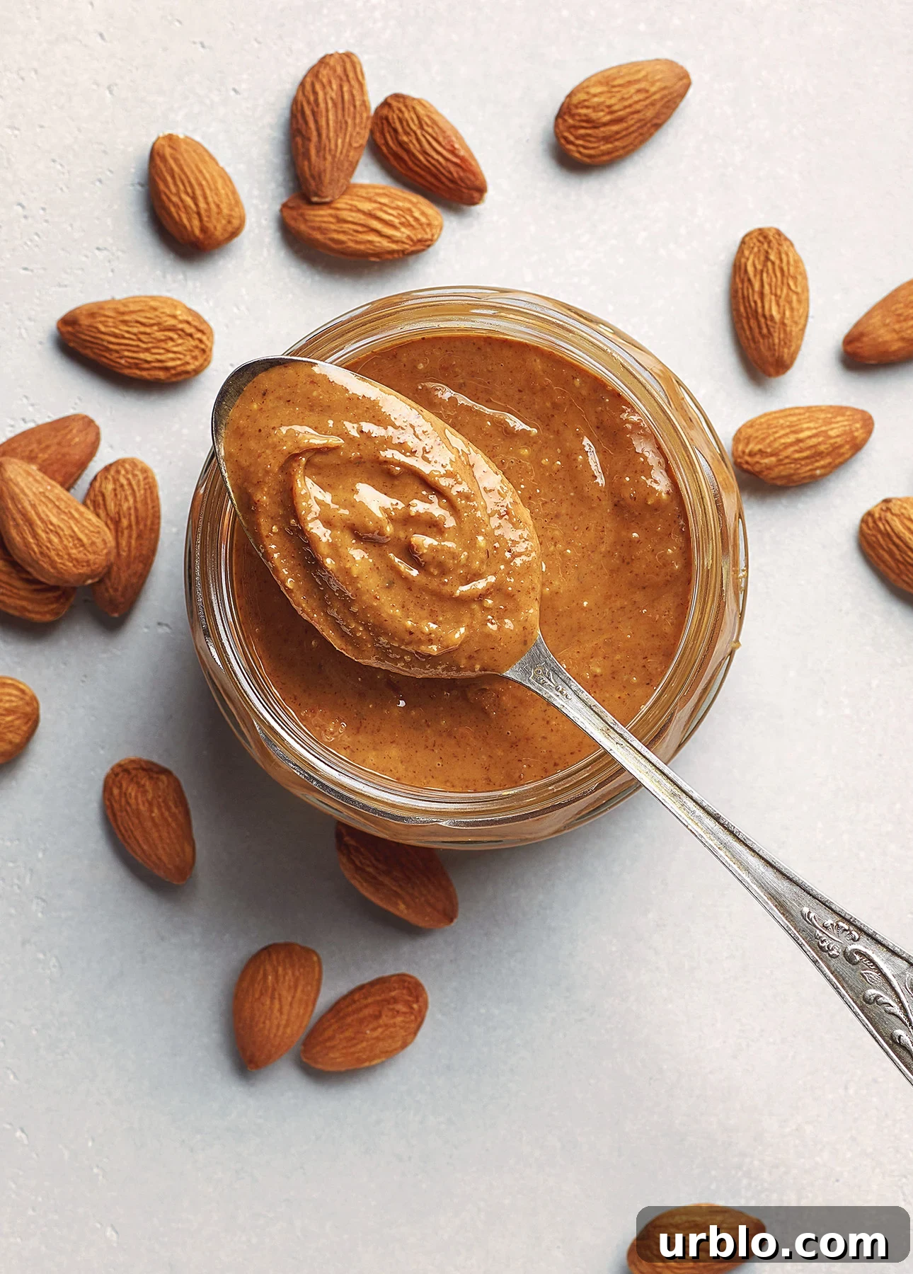 A spoonful of almond butter is held above an open jar filled with the spread. Raw almonds are scattered around the jar on a white surface. // FoodNouveau.com