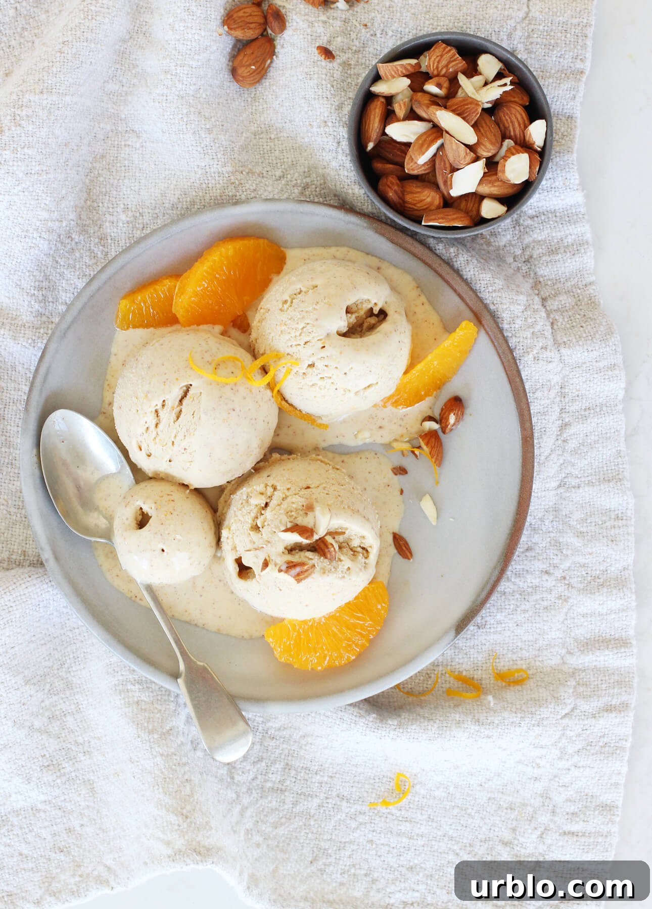 A bowl of orange almond gelato is garnished with almond pieces and fresh orange slices, elegantly placed on a textured napkin. Nearby, a small bowl holds mixed almonds, while a spoon rests invitingly on the plate. // FoodNouveau.com