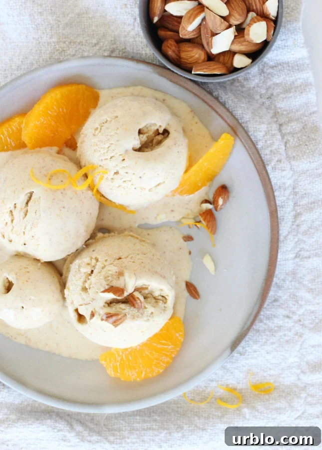 Scoops of orange almond gelato are garnished with sliced almonds and orange segments on a plate. A small bowl filled with almonds is placed beside the plate, all resting on a textured beige cloth. // FoodNouveau.com