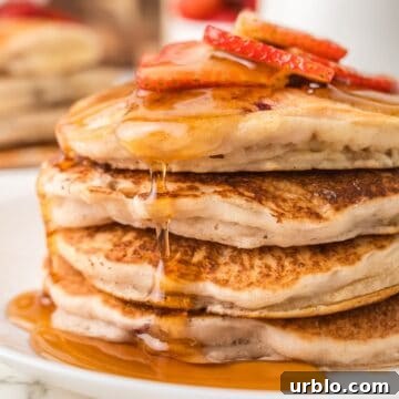 Four pancakes stacked on top of each other with syrup dripped down the sides and sliced strawberries on top.