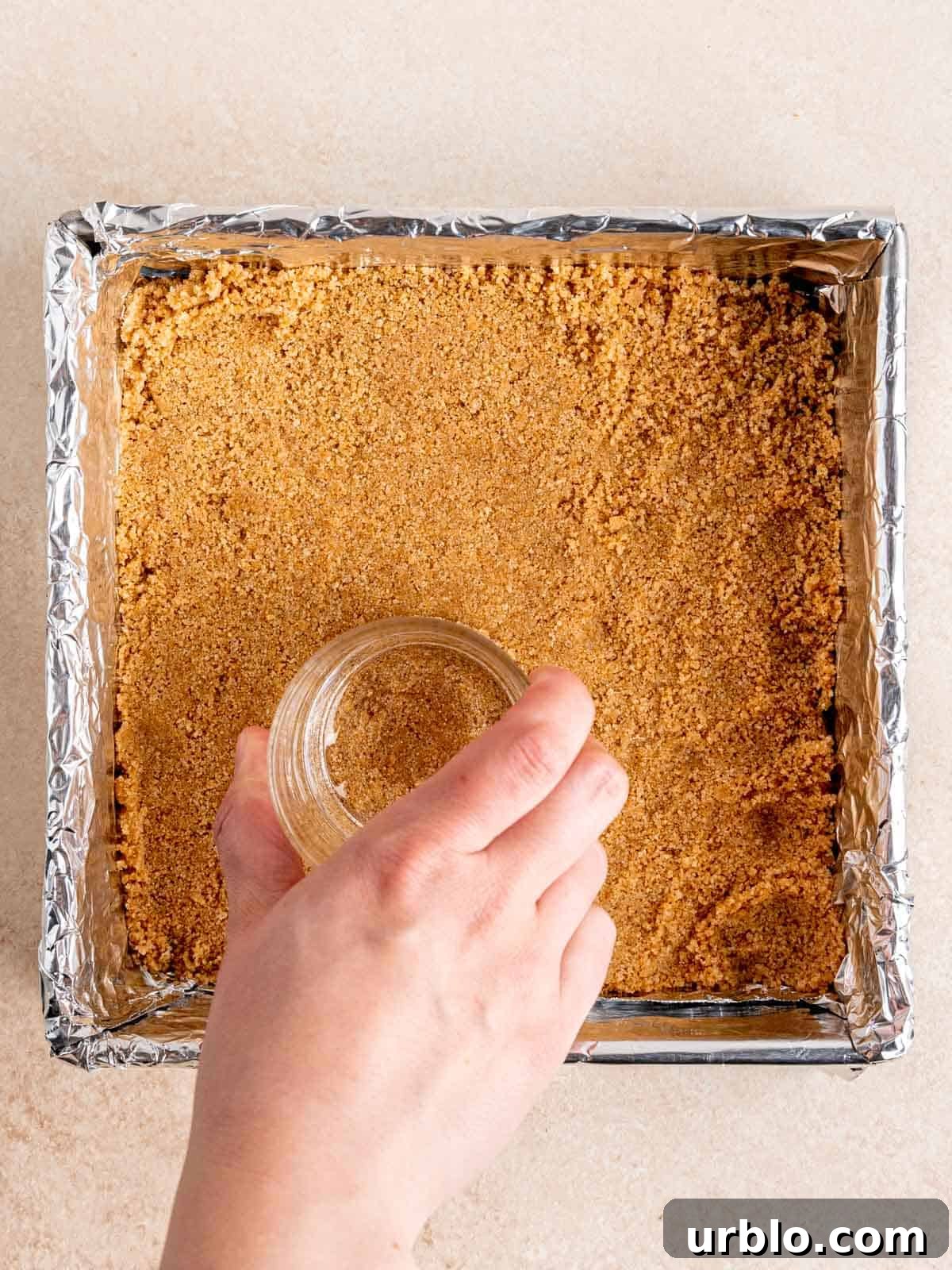 Using the bottom of a glass to press graham cracker crust into a foil-lined pan.