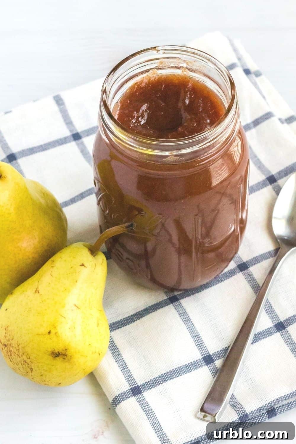 a jar of pear butter on a blue and white checked napkin, with a spoon and two fresh pears next to it.