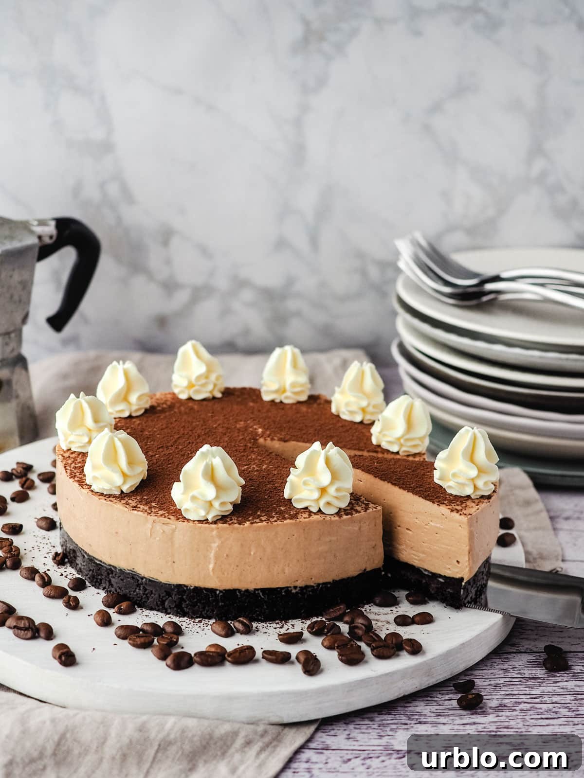 Coffee cheesecake with a slice being taking out of it, with a stack of plates and forks and an espresso maker in the background.