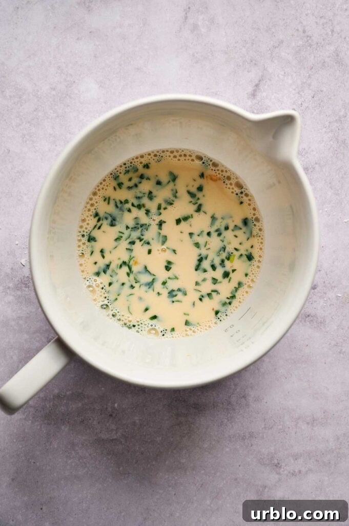 A white mixing bowl containing a beaten egg mixture with chopped herbs, perfect for preparing Air Fryer Chicken Fritters, sits on a light gray surface.
