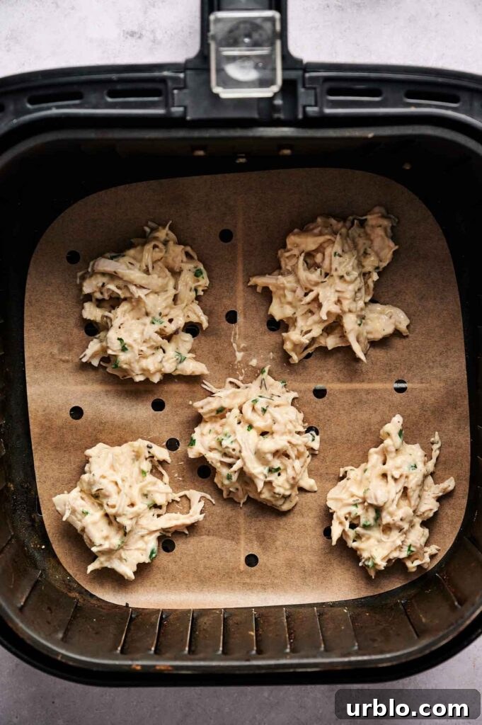 Five raw Air Fryer Chicken Fritters, made with shredded ingredients and herbs, are placed on parchment paper inside an air fryer basket, ready to be cooked.
