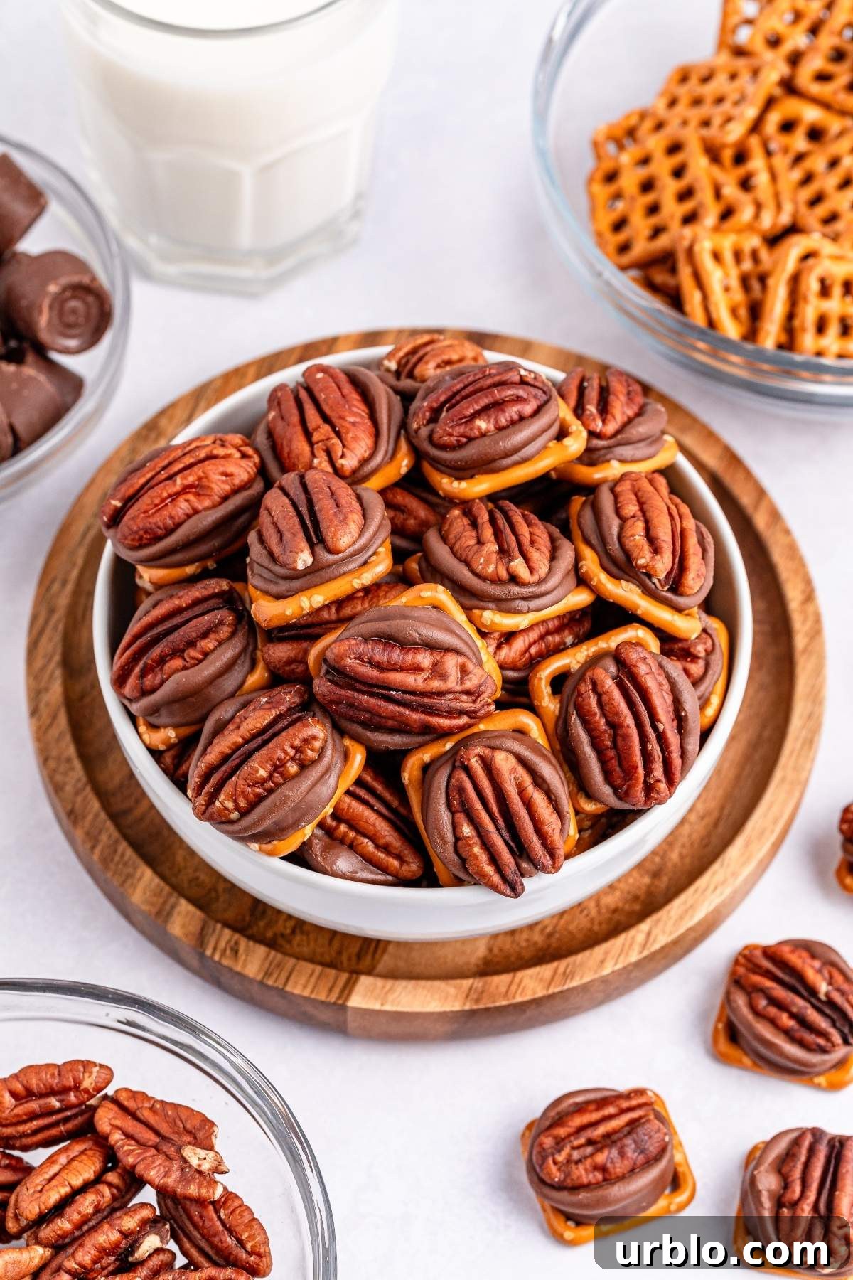 A bowl of chocolate pretzel Rolo turtle candy bites is surrounded by glasses of milk, chocolate candies, pecans, and pretzels on a white surface.