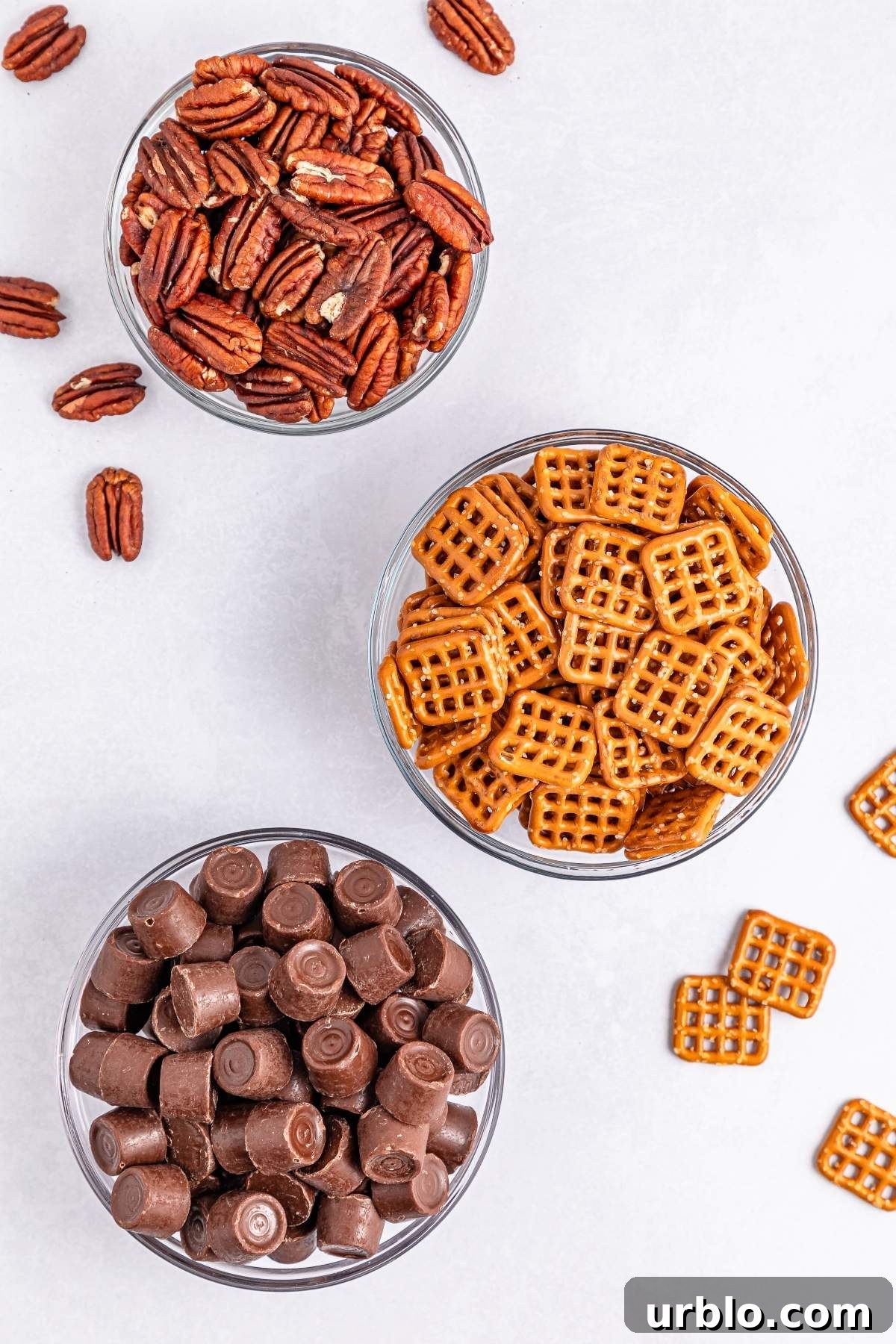 Three bowls filled with pecans, square pretzels, and chocolate candies are arranged on a light surface, creating the perfect setup for making pretzel turtle bites. Pieces are scattered around, adding to the delightful chaos.