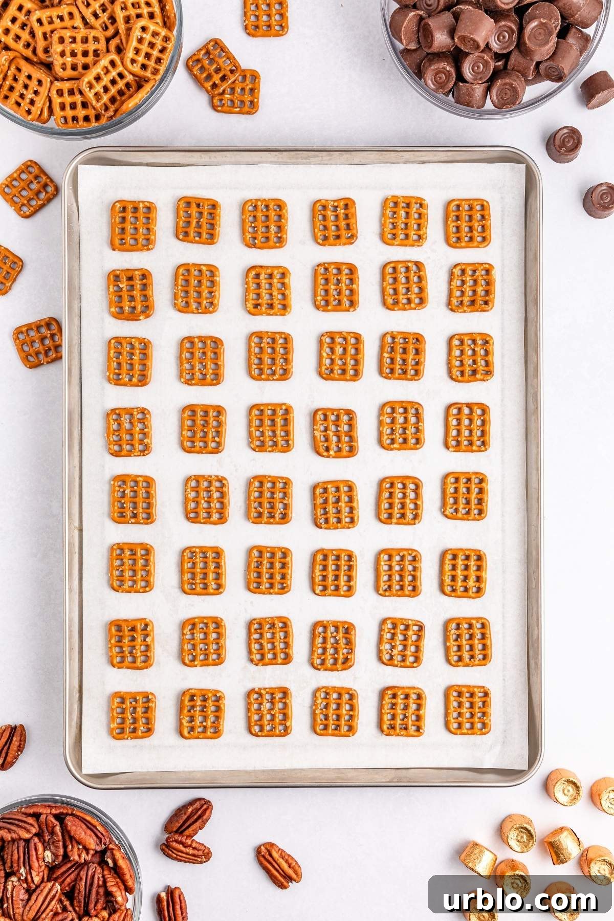 A baking sheet with evenly spaced square pretzels is surrounded by bowls of pecans, chocolate, and additional pretzels on a white surface, ready to become delicious pretzel turtle bites.