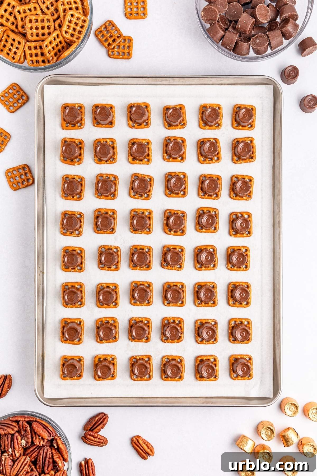 A baking sheet displays pretzel turtle bites, each topped with rich chocolate and nestled pecans. Surrounding bowls brim with pretzels, chocolates, and more pecans, ready for your next creation.