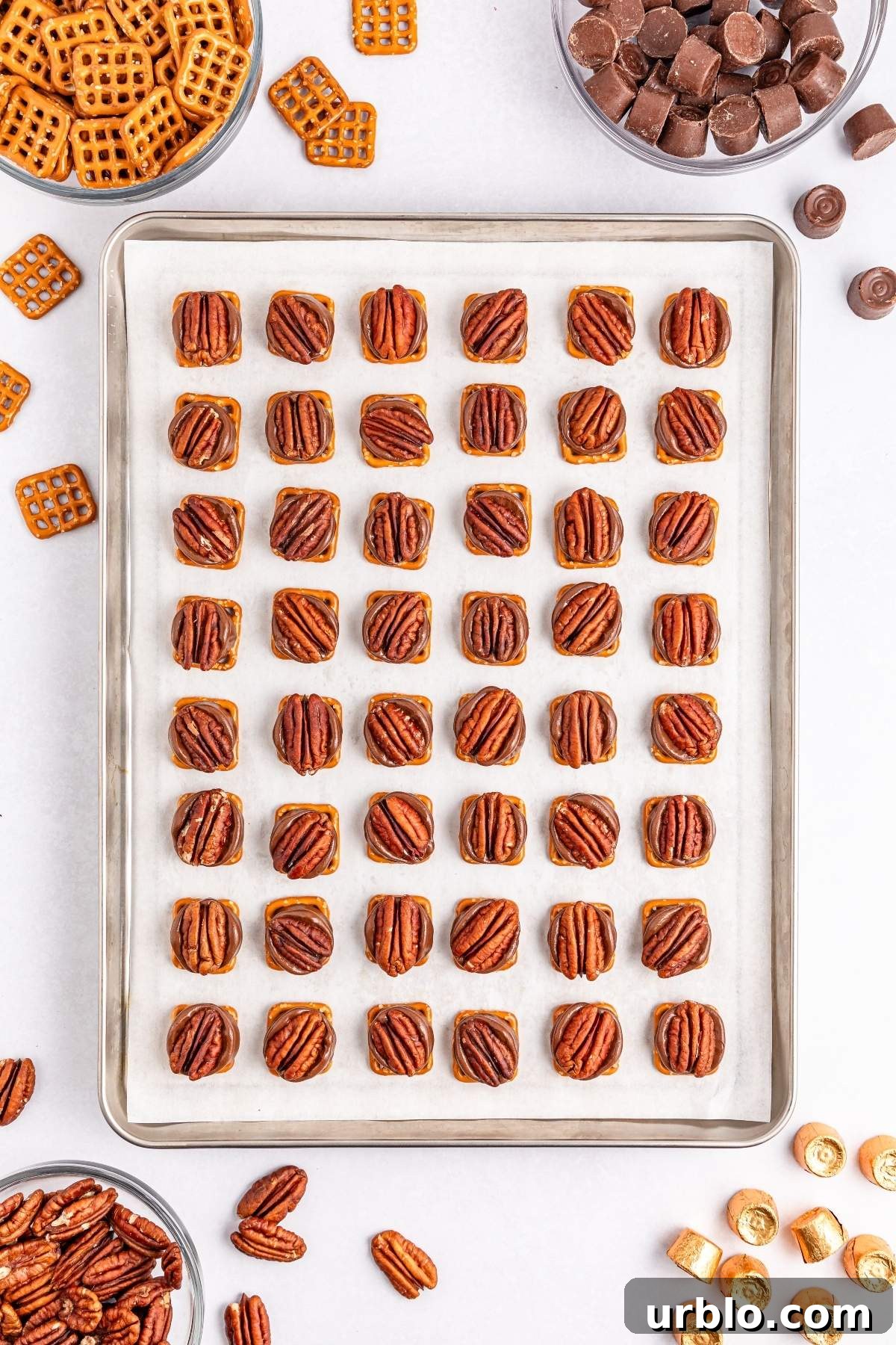 A baking sheet displays pretzel turtle bites, perfectly arranged and topped with pecans. Nearby are bowls filled with pretzels, chocolate pieces, and loose candies.