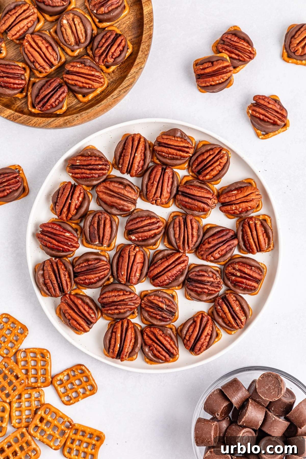 A plate of pretzel turtle bites is topped with pecans and surrounded by extra candies, pretzels, and unwrapped chocolate pieces.