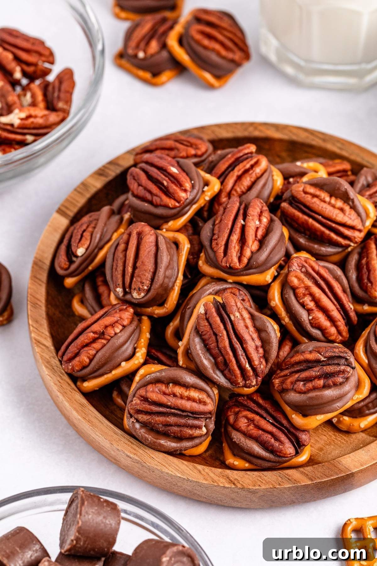 A wooden bowl filled with pretzel Rolo turtle bites topped with caramel, melted chocolate, and pecans. Additional bowls of similar snacks and a glass of milk are in the background.