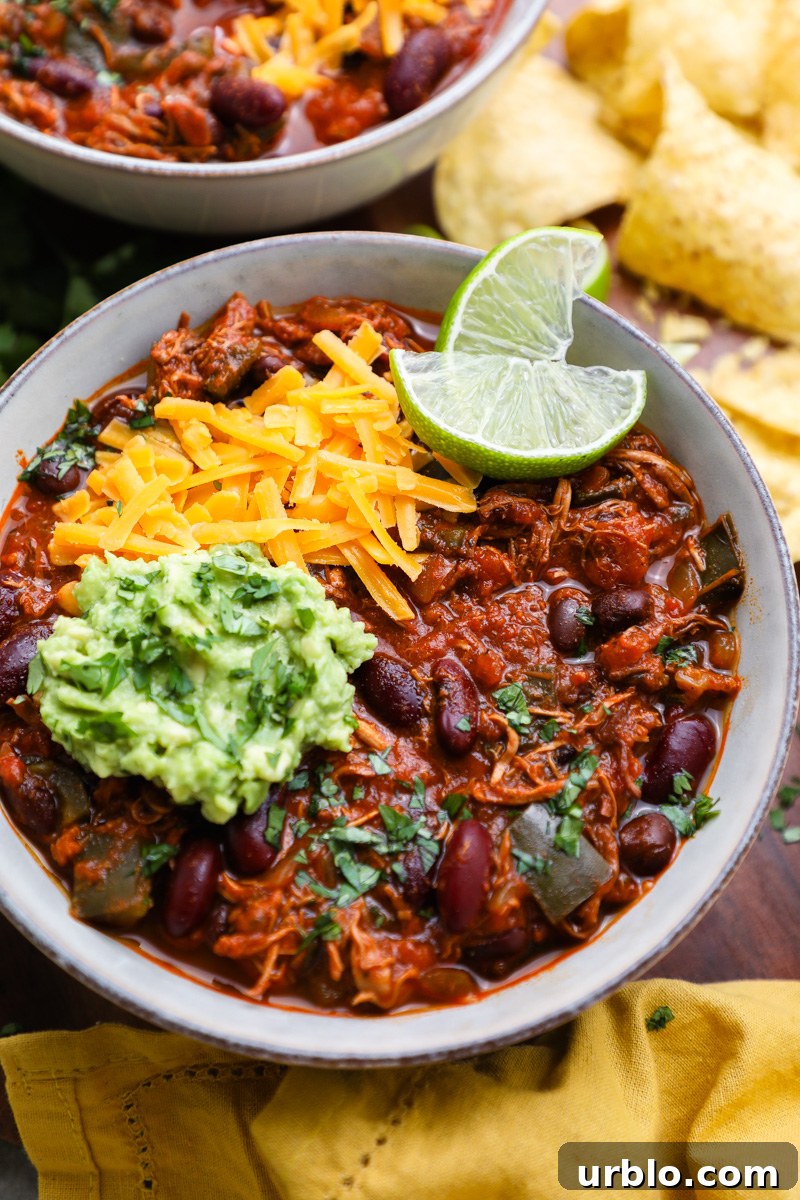 A bowl of crockpot chipotle chicken chili topped with chopped cilantro, shredded cheese and avocado sitting next to another bowl of chipotle chicken chili