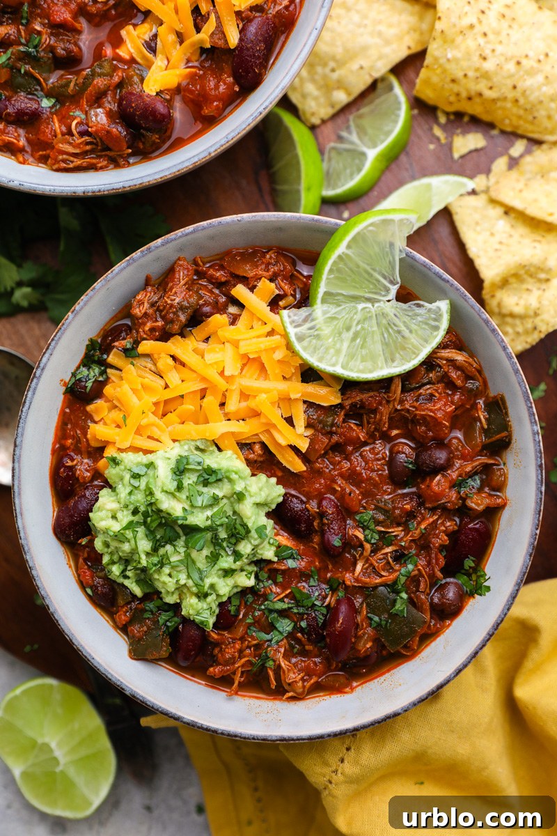 A serving bowl of chipotle chicken chili topped with mashed avocado, shredded cheese and fresh lime wedges