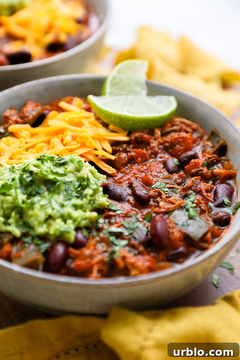 A side view of a bowl of chipotle chicken chili topped with shredded cheese surrounded by tortilla chips