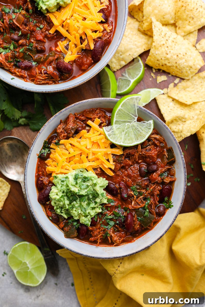 2 bowls of crockpot chipotle chicken chili sitting on a serving tray surrounded by tortilla chips, lime wedges and fresh cilantro