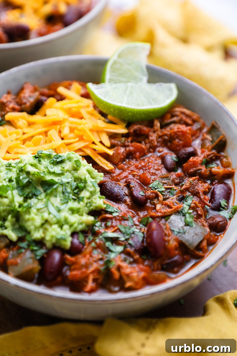 A small serving bowl of chipotle chicken chili topped with shredded cheese, mashed avocado, chopped cilantro and fresh lime wedges