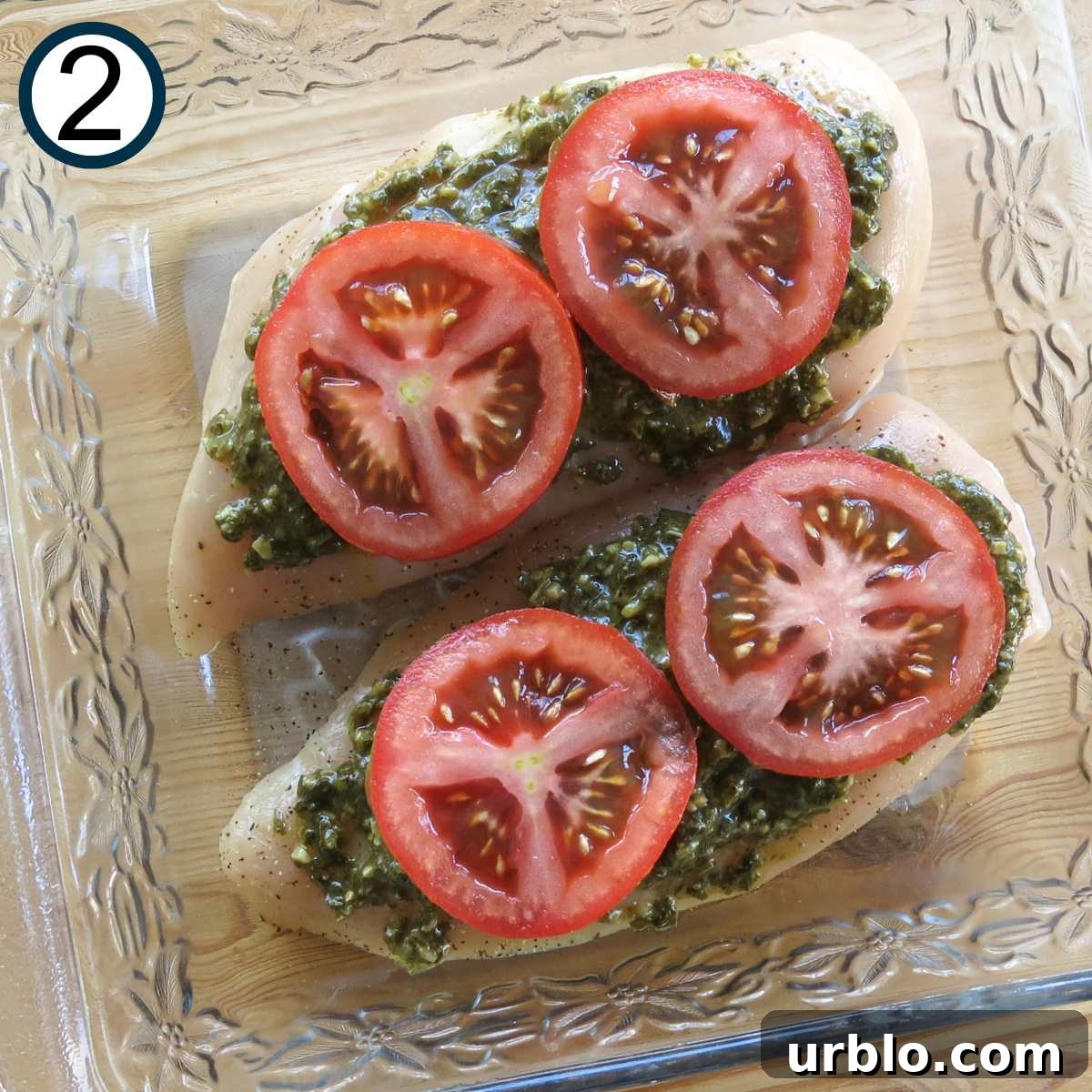 Crispy Baked Pesto Chicken Breast with Parmesan Crust 4 Slices of tomato on top of two boneless, skinless chicken breasts with pesto spread on top in a glass baking dish.
