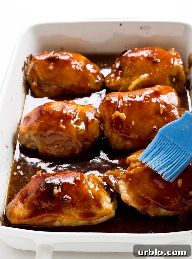 Baked Honey Garlic Chicken Thighs in a white baking dish being brushed with the honey garlic sauce 