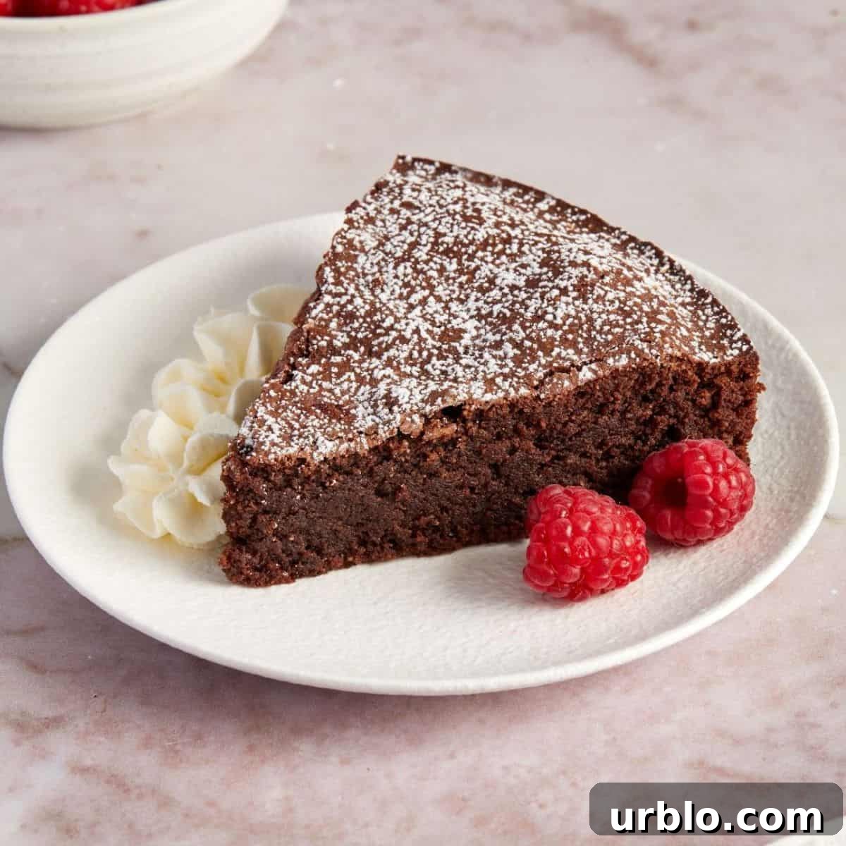 Slice of Torta Caprese, on a small round white plate, with some raspberries and fresh cream on the side.