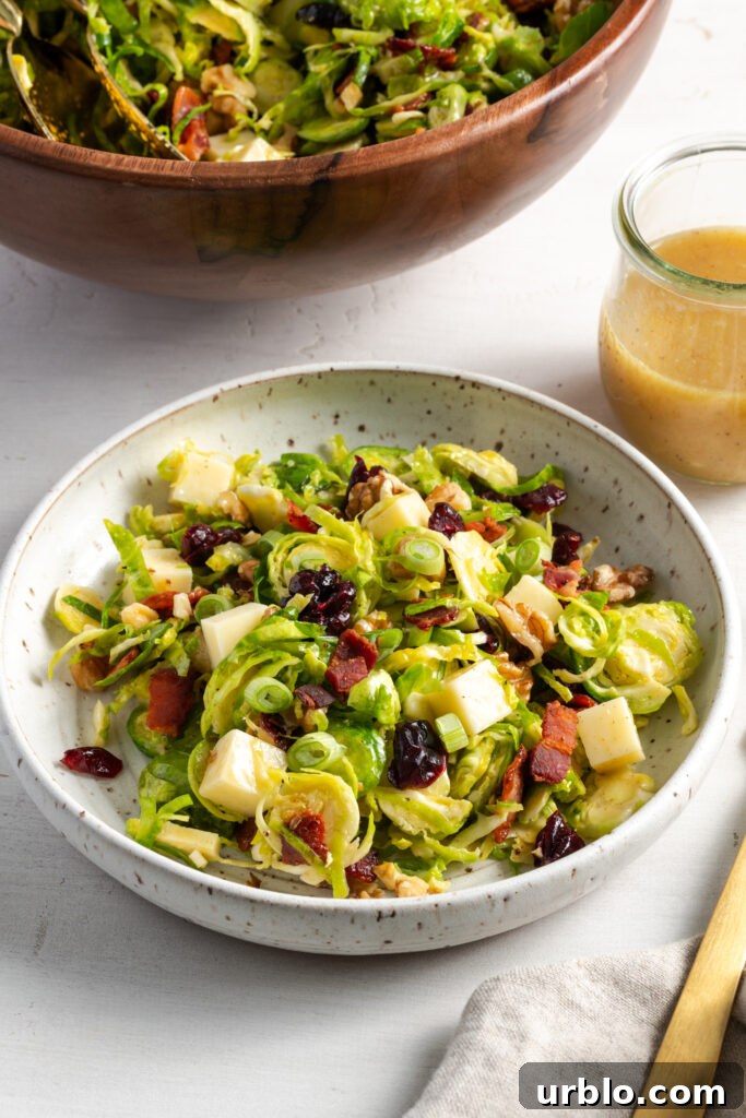45 degree angle shot of a white flecked earthenware shallow bowl filled with a serving of shaved brussels sprouts salad on a white table with the wooden serving bowl and a small glass jar with extra vinaigrette.
