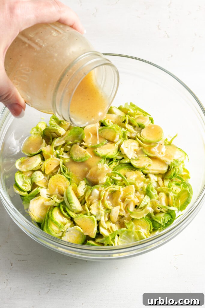 hand pouring vinaigrette from a mason jar over the shaved brussels sprouts in the bowl.