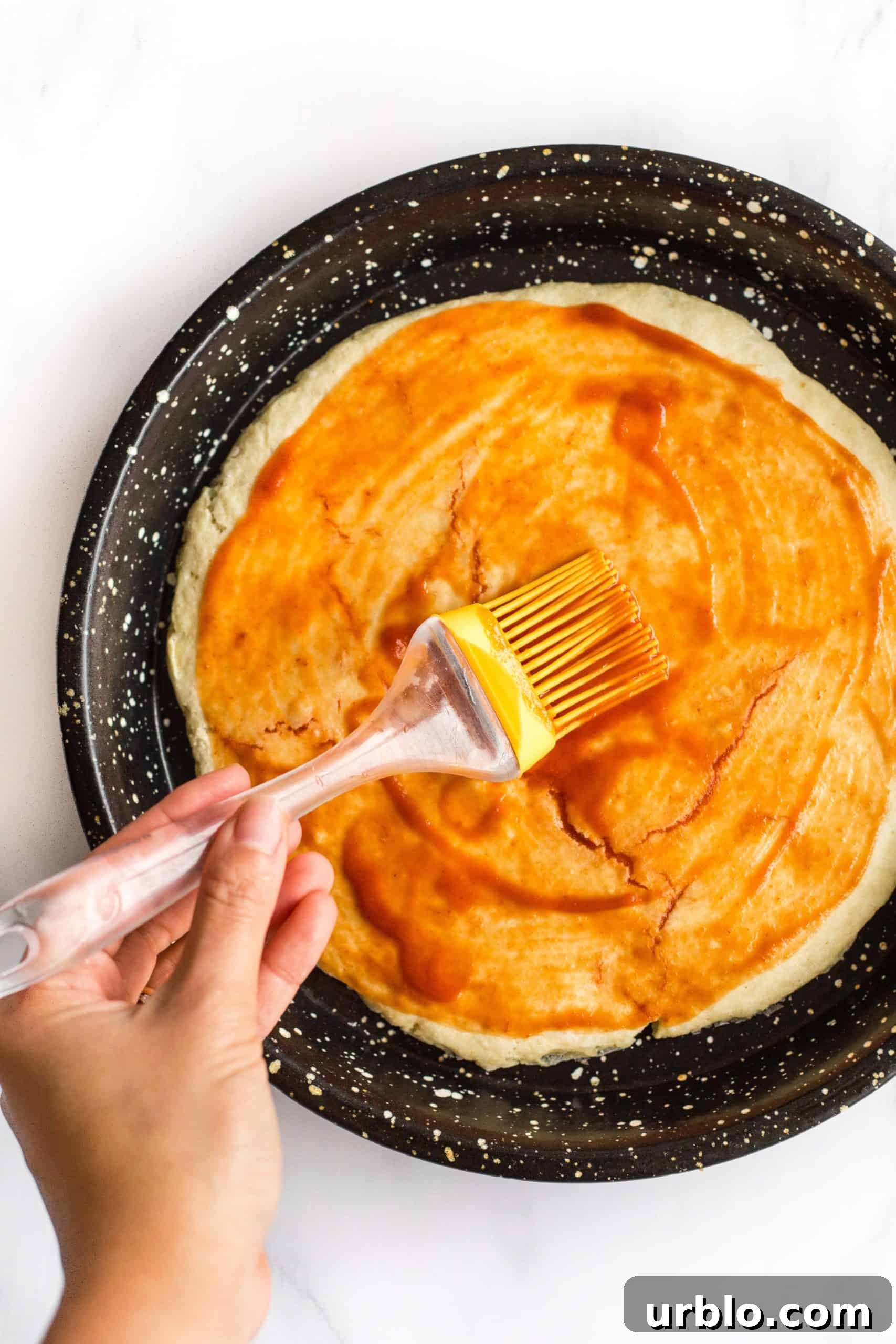 Using a silicon brush to spread tomato sauce over a pre-baked pizza crust.