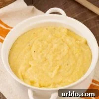 A round, white casserole of creamy polenta atop a striped tea towel with a wooden spoon to the rear.