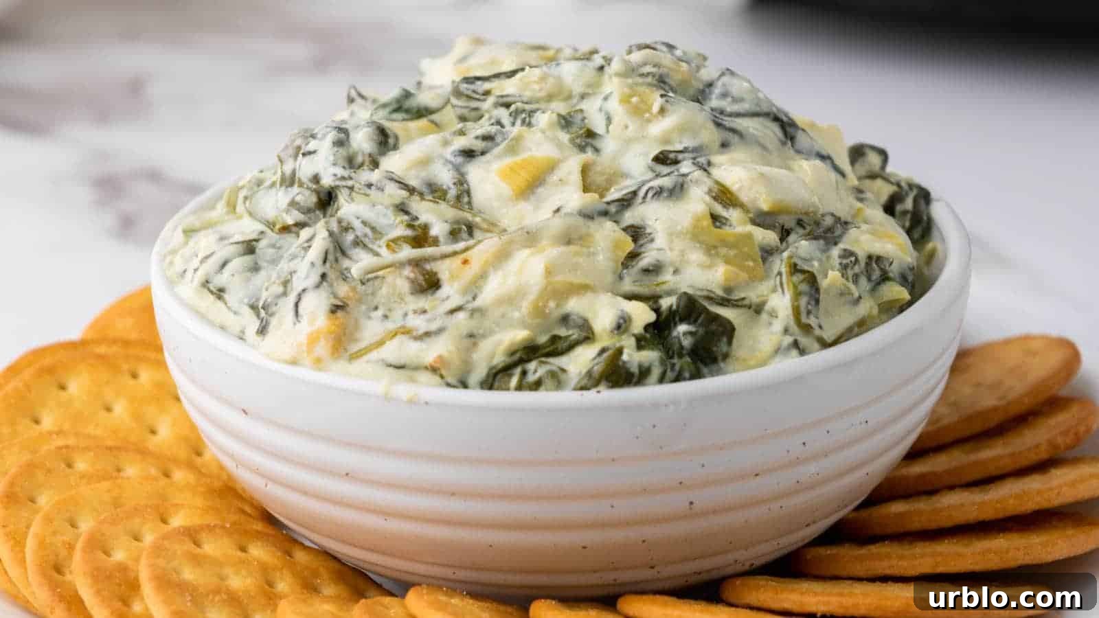 21 Crowd-Pleasing Potluck Recipes That Outshine Store-Bought Dishes 3 A white bowl of creamy spinach artichoke dip surrounded by round crackers on a marble surface.