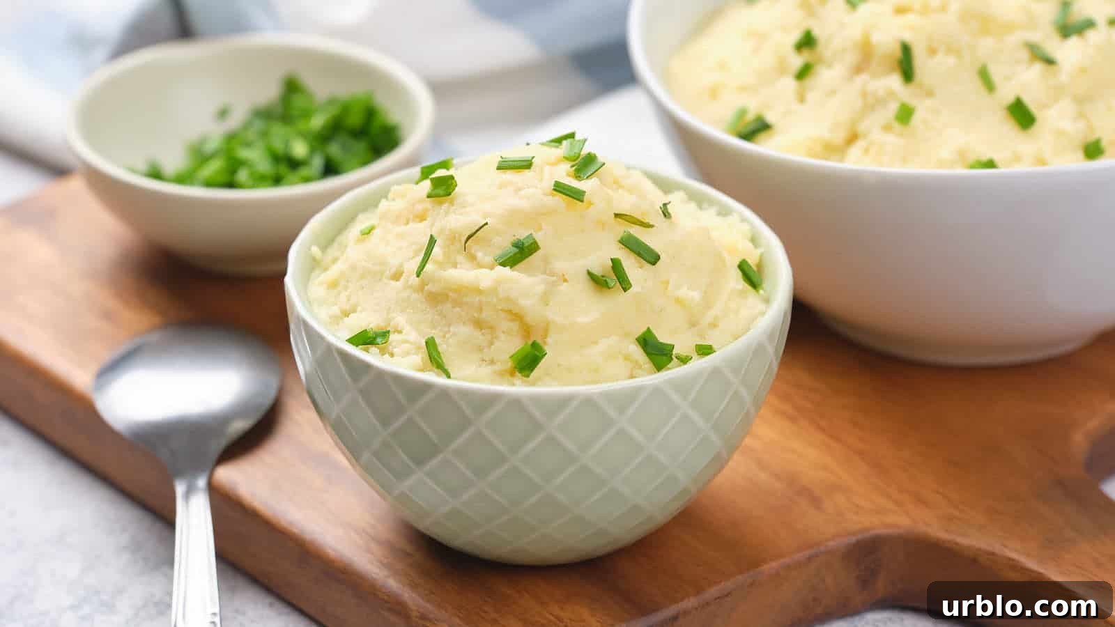 21 Crowd-Pleasing Potluck Recipes That Outshine Store-Bought Dishes 5 A bowl of mashed potatoes with chopped chives on a wooden board, accompanied by a spoon and a small dish of chives.