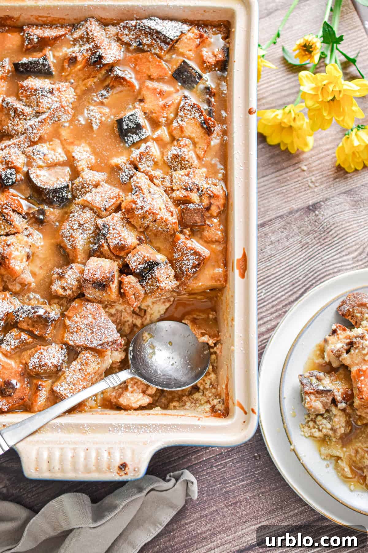 A spoon is in a pan of sourdough bread pudding with the plate of sourdough bread pudding next to it.