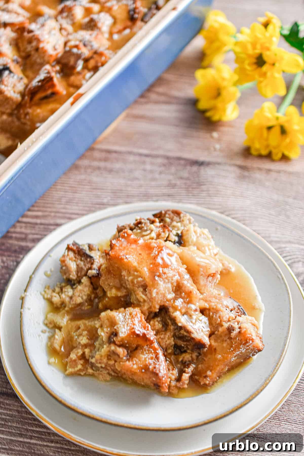 A portion of the sourdough bread pudding is on a plate with yellow flowers in the back and the blue pan behind it.