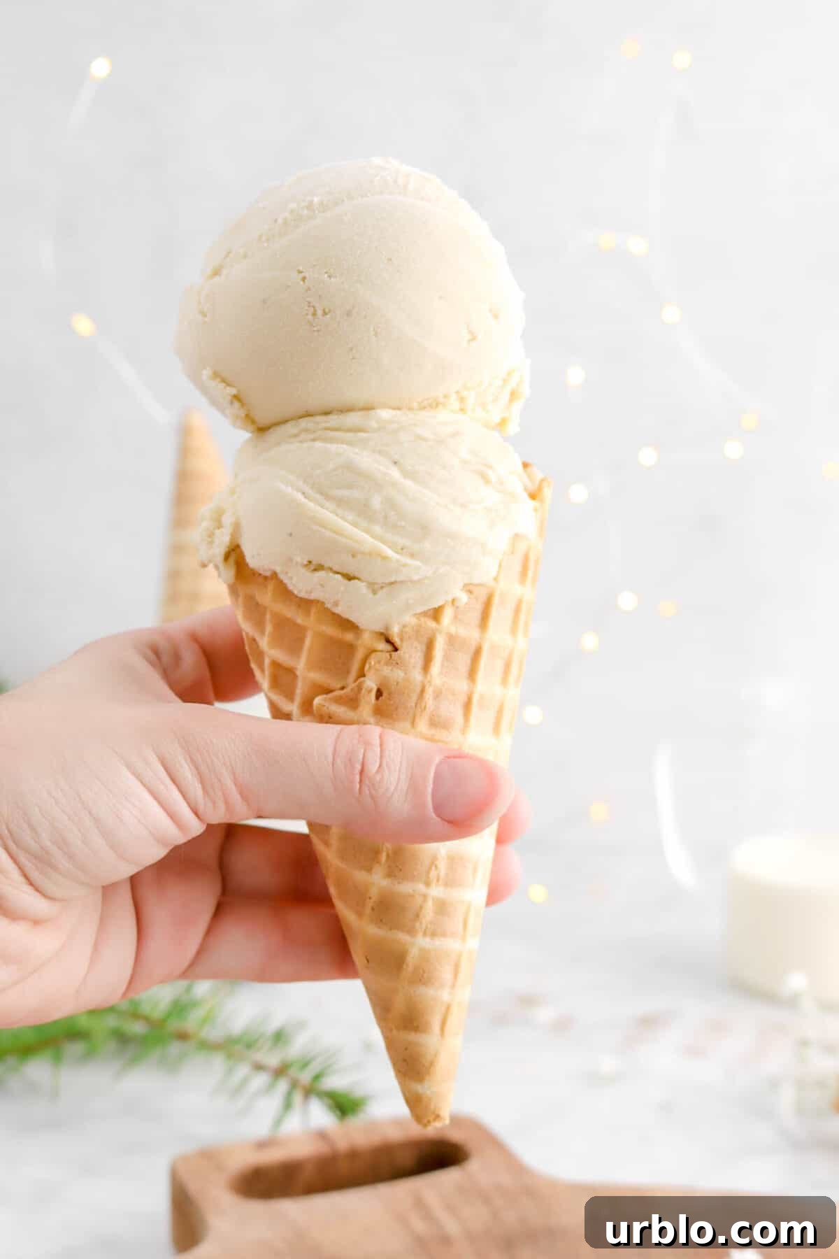 hand holding ice cream cone with two scoops of eggnog ice cream with fairy lights behind