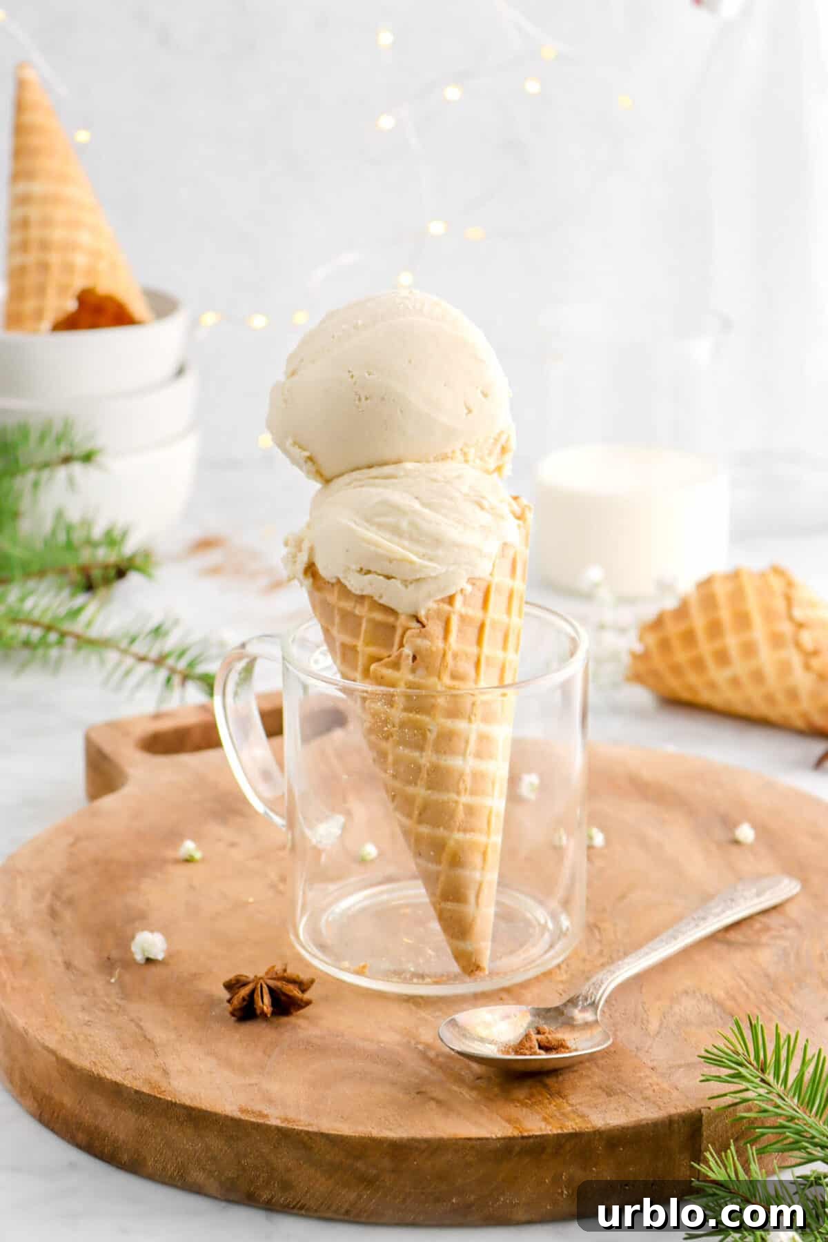 two scoops of eggnog ice cream in cone with glass mug on wood board with spoon, star anise pod, mug of eggnog behind, and pine branches