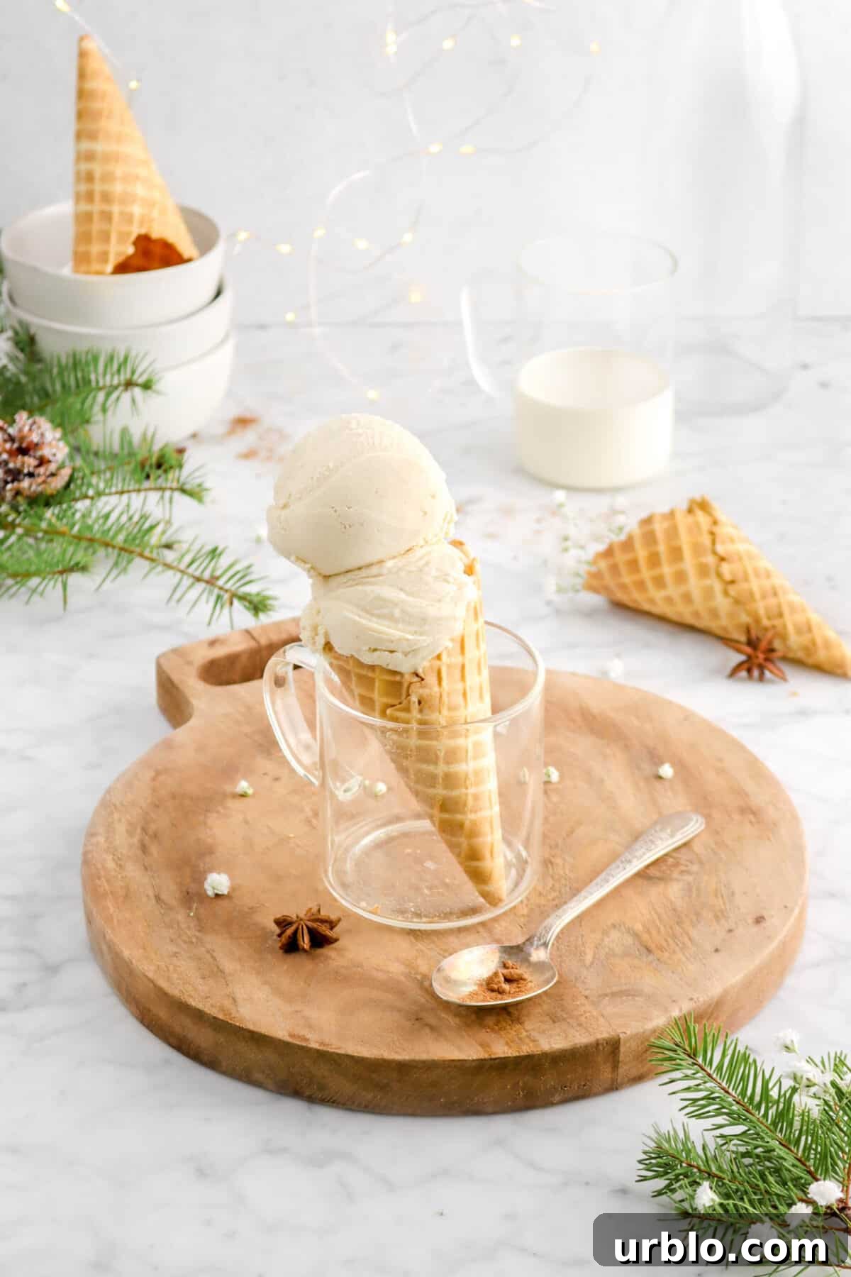 eggnog ice cream in waffle cone in glass mug on wood serving board with spoon, star anise pod, flowers, pine branches, two empty ice cream cones, a mug of eggnog, and fairy lights
