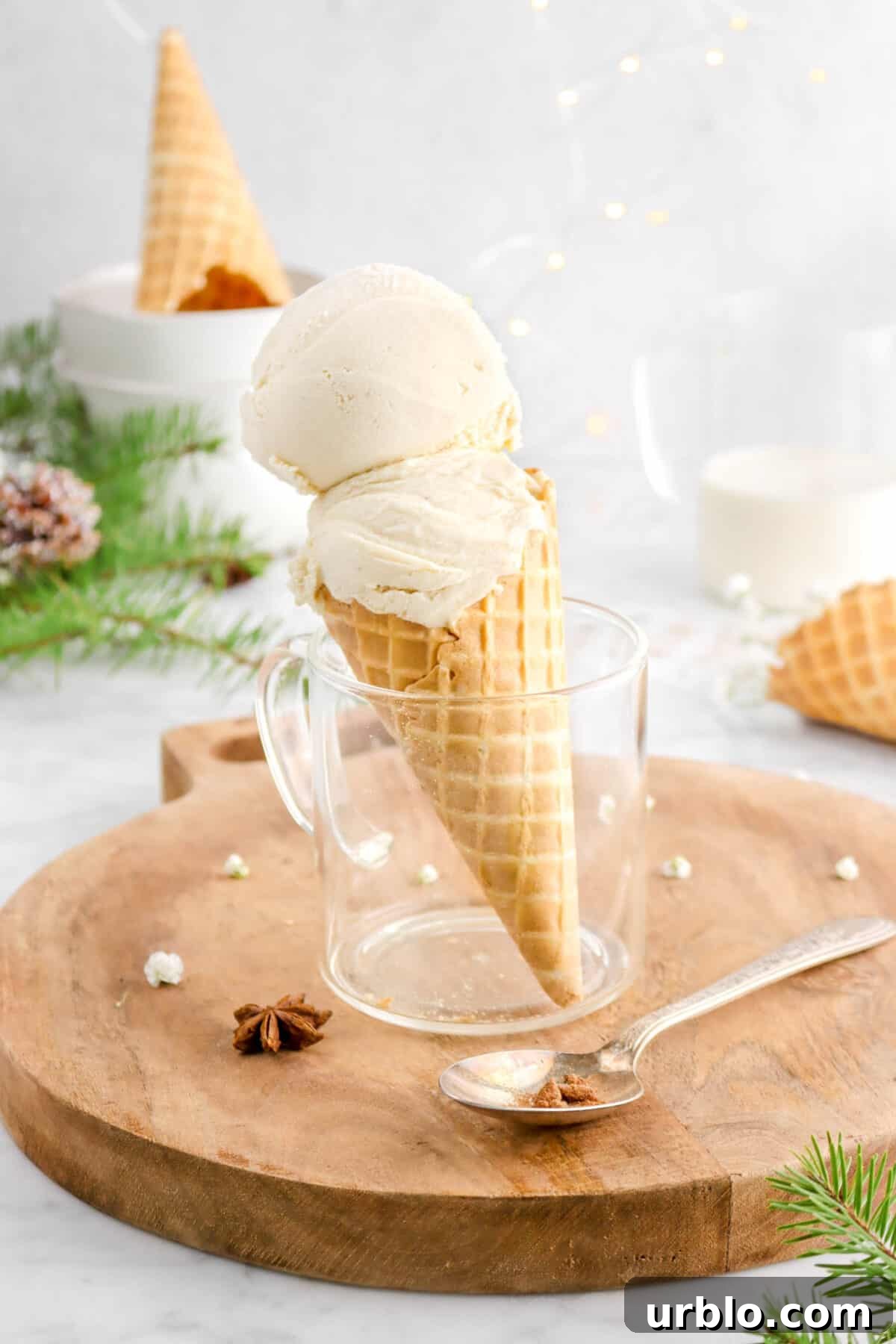 two scoops of ice cream in waffle cone in glass mug with spoon beside, upside down cone behind in stack of bowls, pine branch, and fairy lights