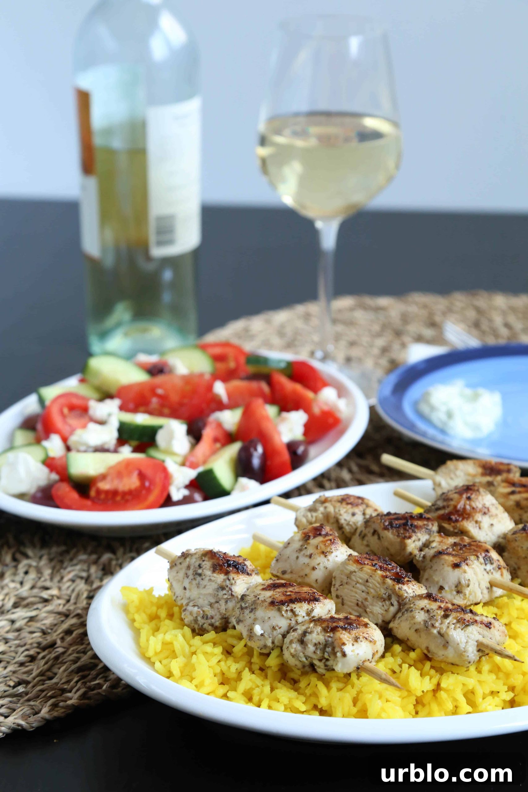 Chicken kabobs, rice, Greek salad, wine glasses, and a bottle of wine on a dinner table.