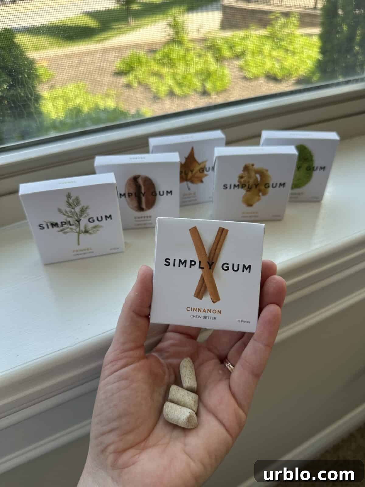 A person showcasing a Cinnamon-flavored gum from Simply Gum, with five other boxes in the background.