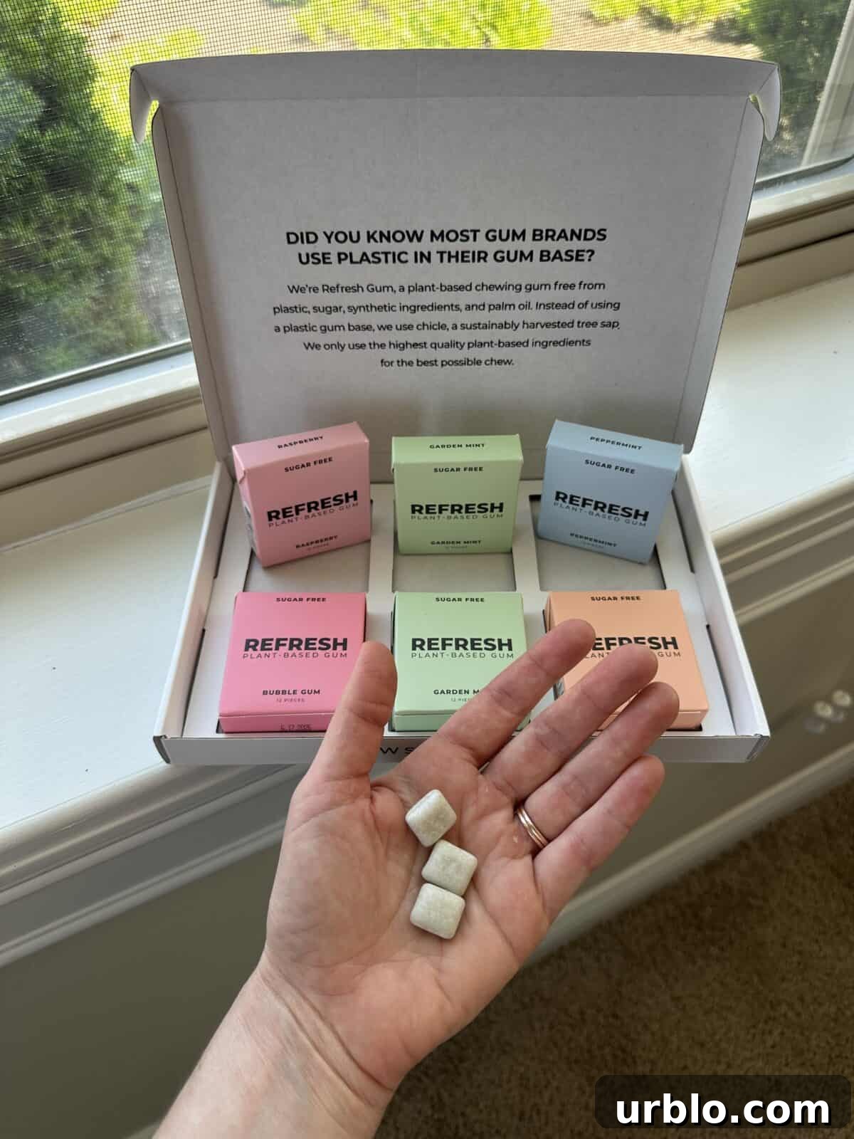 A package of plastic-free gum from Refresh, containing six boxes inside, with a hand showing three pieces of gum.