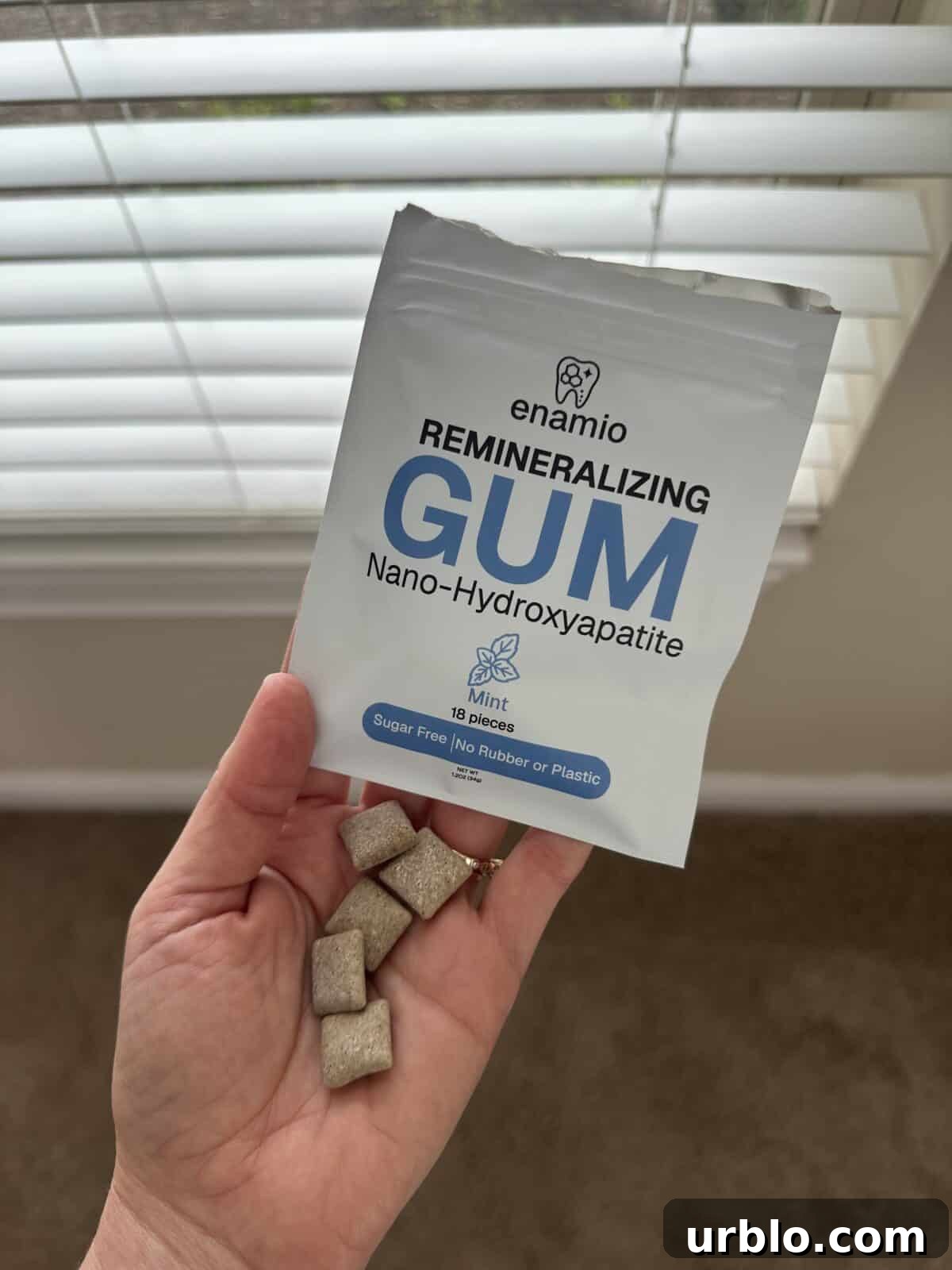 A hand holding several square pieces of beige chewing gum in front of a white package labeled “enamio Remineralizing Gum – Nano-Hydroxyapatite, Mint, 18 pieces, Sugar Free, No Rubber or Plastic.”