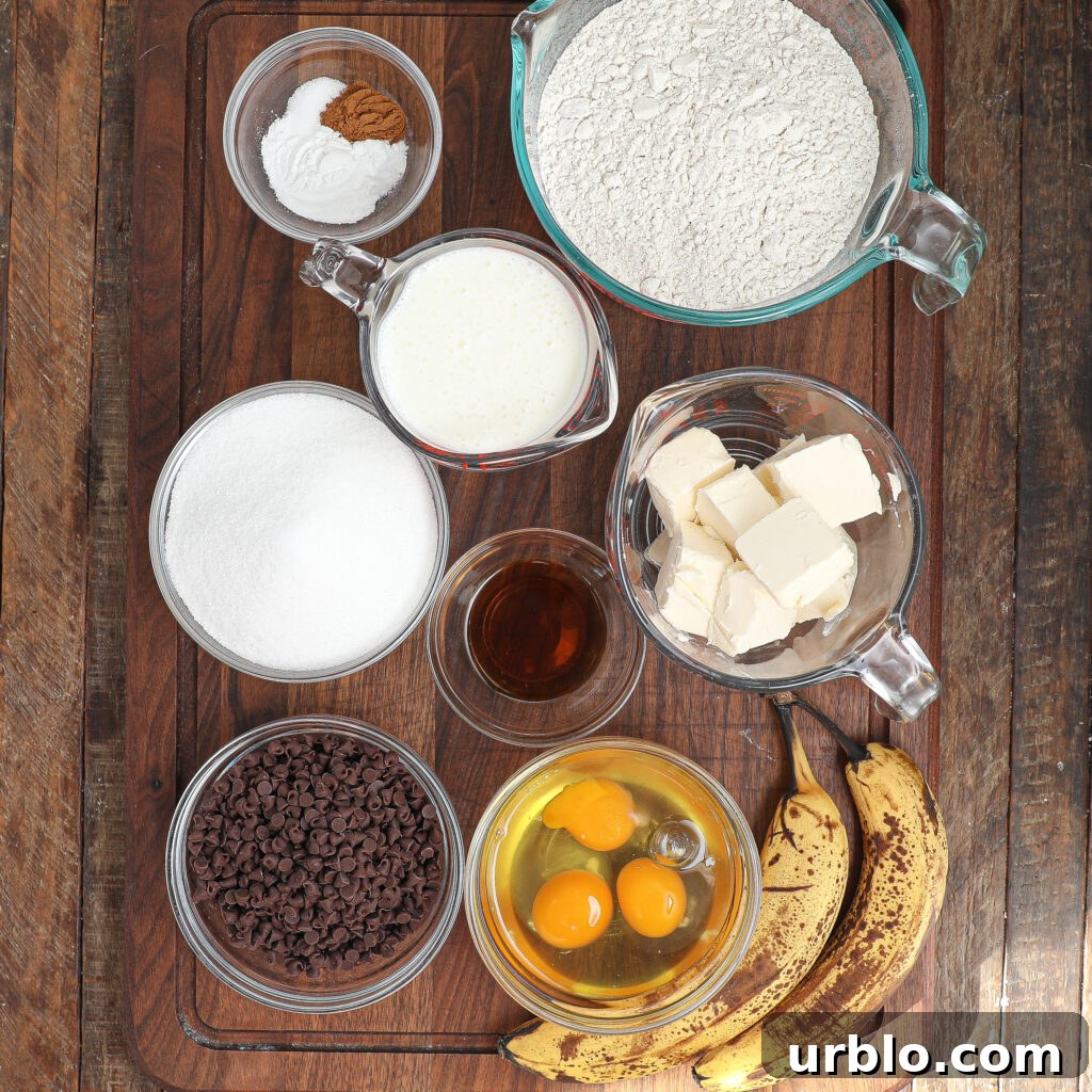 A wooden surface with bowls and measuring cups holding ingredients for a Banana Chocolate Chip Snack Cake: flour, sugar, butter, chocolate chips, eggs, vanilla, buttermilk, cinnamon, baking soda, and two ripe bananas—all neatly arranged for baking.