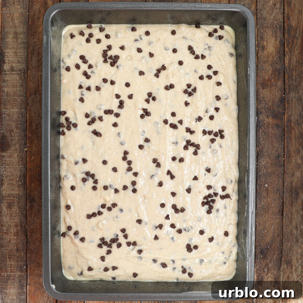 A rectangular metal baking pan filled with raw Banana Chocolate Chip Snack Cake dough, topped with chocolate chips, sits on a rustic wooden surface.