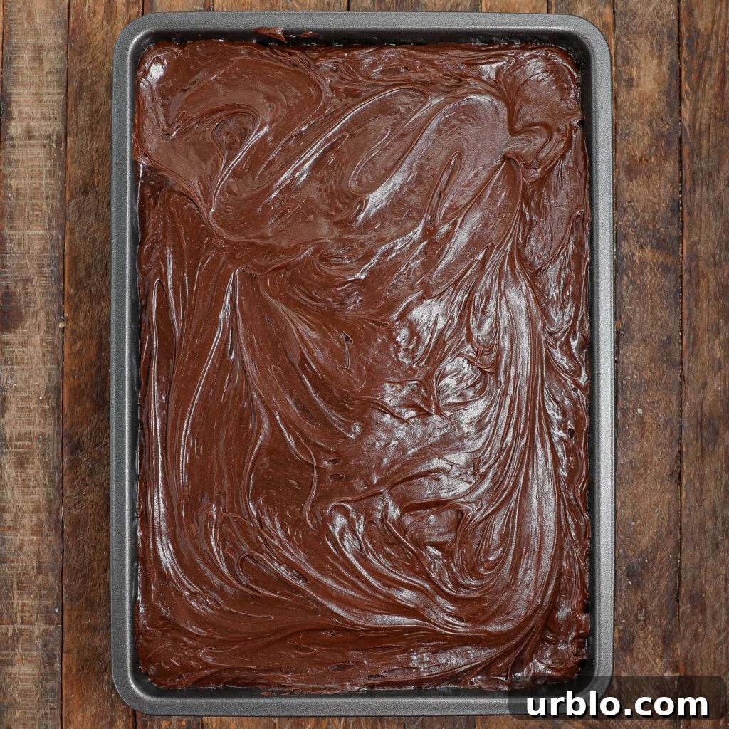 A baking pan filled with rich, glossy chocolate frosting swirled smoothly over a Banana Chocolate Chip Snack Cake, set on a rustic wooden surface.