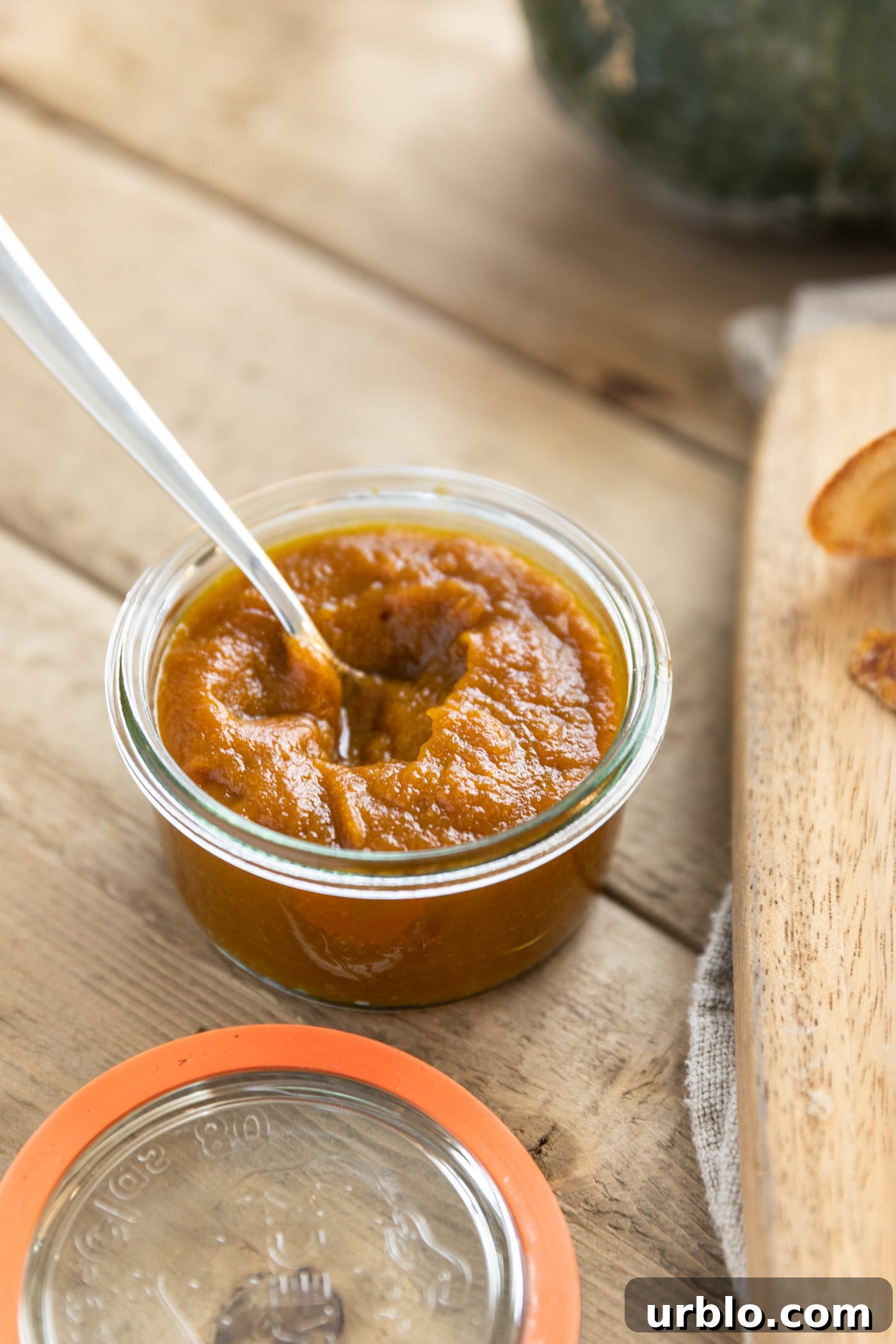 Glass jar holding brown colored spread with spoon in mixture on top of wood table with pumpkin in background
